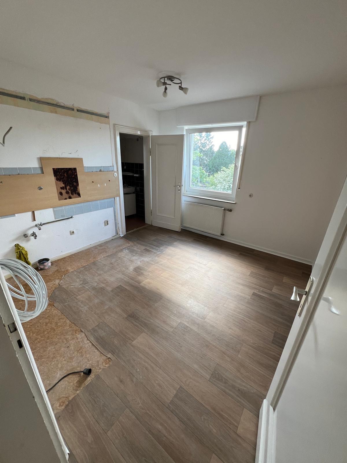 Ein leerer Raum mit braunem Laminatboden und großem Fenster, helle Wände.